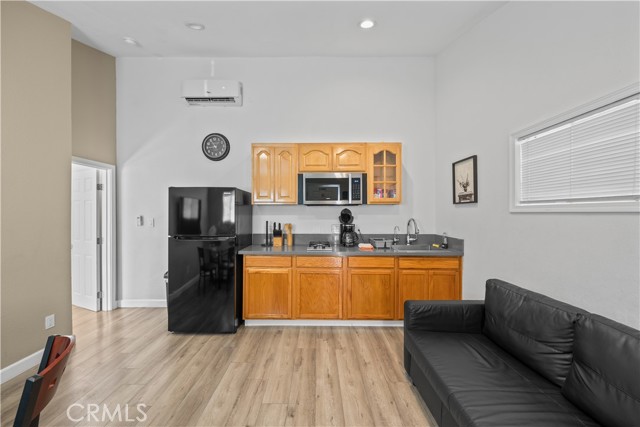 A small kitchenette featuring light wood cabinets, a black refrigerator, and an adjacent living area with a black leather sofa.
