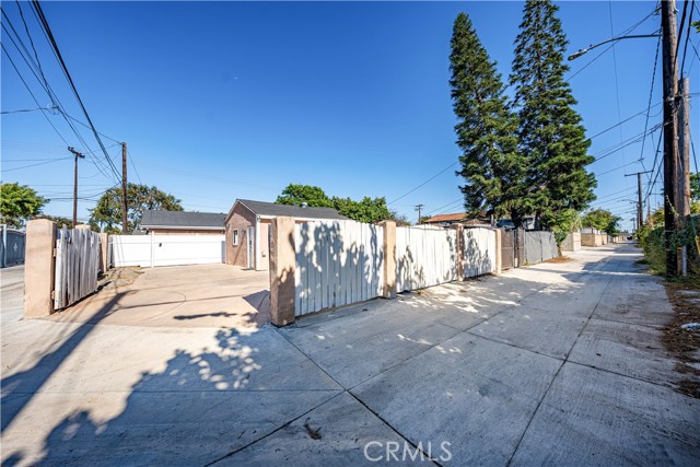 A rear view of a property showing a concrete driveway, a white wooden fence, and tall pine trees under a clear blue sky, viewed from a narrow paved alleyway.