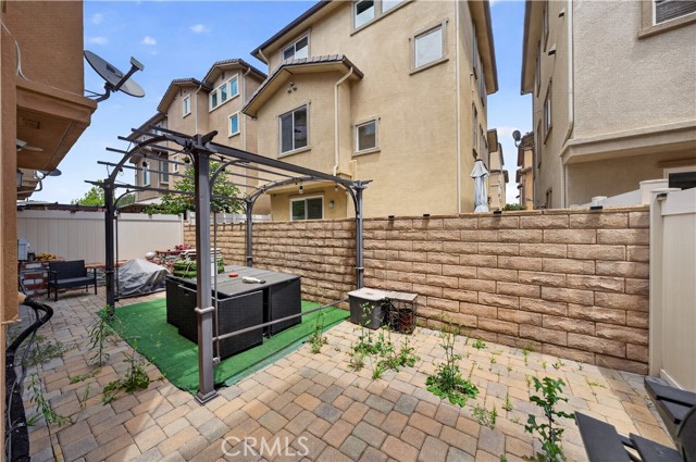 A small backyard patio featuring a metal gazebo structure covering a dining set and synthetic grass, with a stone paver floor and masonry wall.