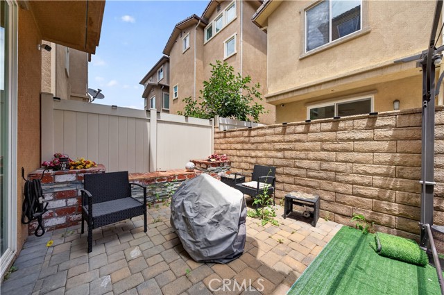 A small paved residential patio featuring outdoor furniture, a grill covered with a tarp, and brick wall accents, enclosed by privacy fencing.
