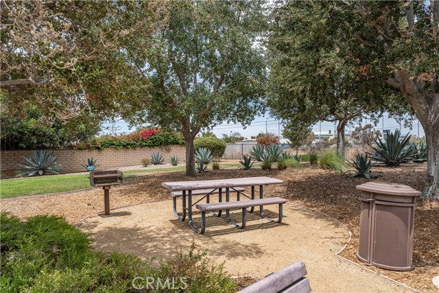 A community picnic area featuring a metal picnic table, a park grill, and a trash receptacle under shade trees