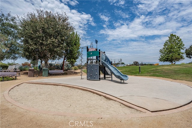 A community playground area with a climbing structure and slide set in a sand pit surrounded by green space and trees.