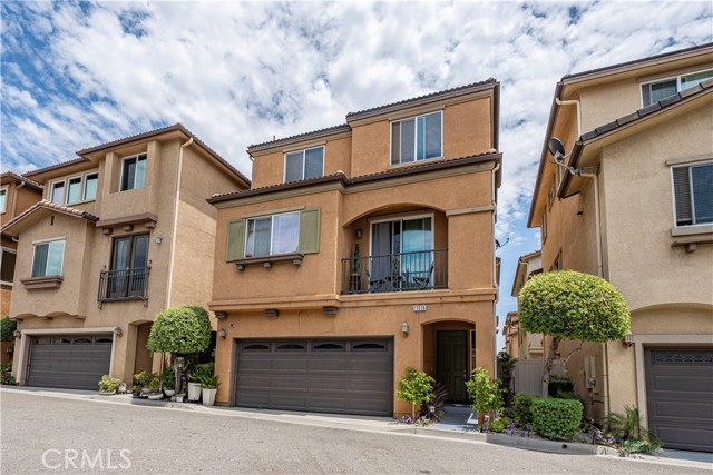 A street-level perspective of a tan, three-story residential townhome with a dark garage door and a second-story balcony.