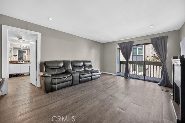 A living room with gray walls, wood-look laminate flooring, a black leather sectional sofa, and sliding glass doors leading to a balcony.