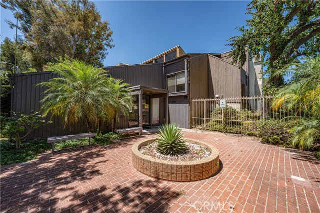 Exterior view of the entrance area of a multi-story building with brown siding, a brick patio, a circular brick planter, palm trees, and metal fencing.