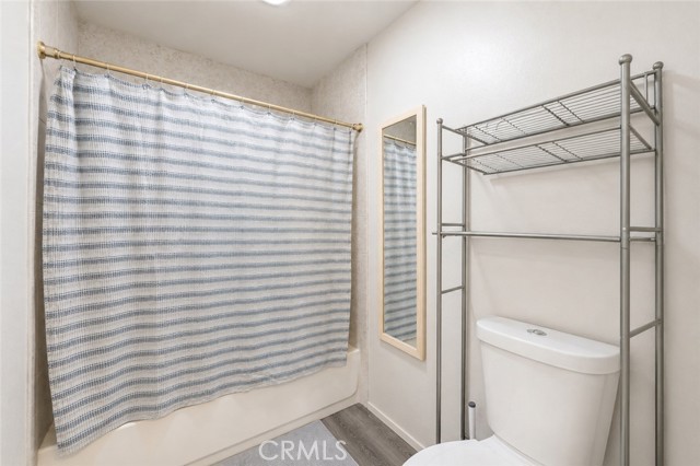 A standard bathroom with a white tub enclosed by a striped blue and white shower curtain, next to a white toilet with a grey metal storage rack above it and a small wall mirror.
