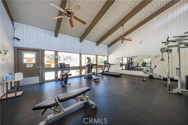 A community fitness center with vaulted ceilings, various gym equipment including a treadmill and weight bench, and large windows looking out towards a pool area.