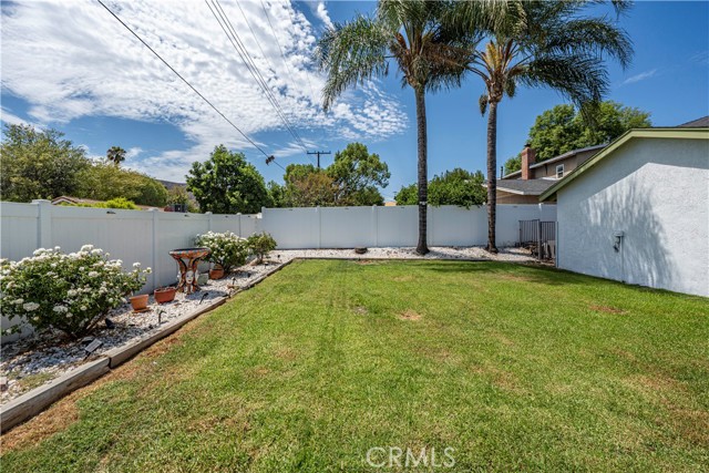 A wide shot of a residential backyard with a green lawn, white vinyl fencing, and palm trees under a blue sky.