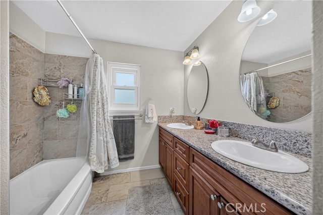 A clean, well-lit bathroom featuring a wooden double vanity with granite countertops, oval mirrors, and a tile-surround tub/shower area.