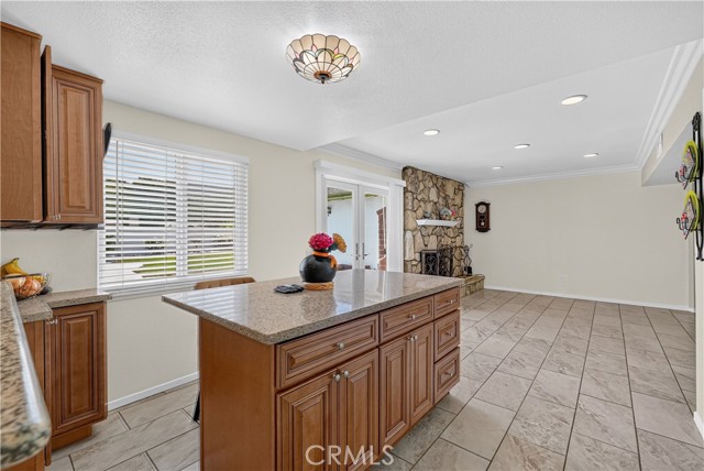 A kitchen with wooden cabinets, a granite topped island, and tile flooring, looking toward a fireplace in the background.