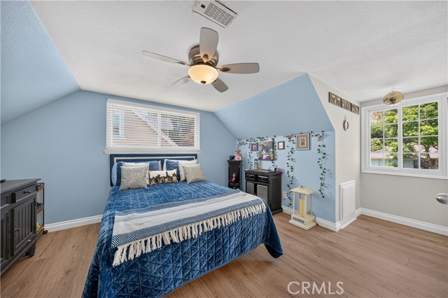 A bedroom with light blue walls, a bed with a blue quilted cover, ceiling fan, and windows on adjacent walls.