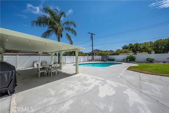 Spacious backyard featuring a large concrete patio with a covered seating area, an outdoor dining set, a swimming pool, and a manicured lawn under a clear blue sky.