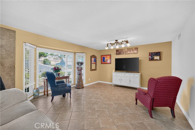 Spacious living room featuring tile flooring, a mounted flat-screen television on a white media console, a bay window with a blue armchair, and a red patterned wingback chair.