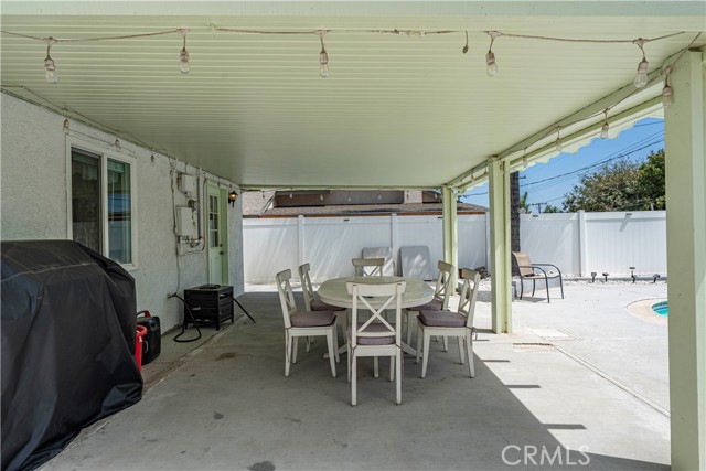 A covered patio area with a dining table and chairs, adjacent to a pool deck with a white vinyl fence in the background.