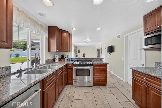 A well-lit kitchen featuring rich wood cabinets, speckled granite countertops, stainless steel appliances, and tile flooring, with a view of the backyard through the window.