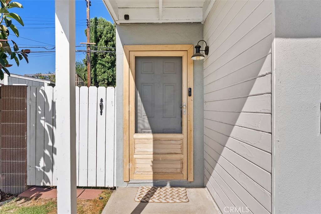 Front entry of 9925 Rutland Avenue with a gray door framed in natural wood, white siding, and a white picket fence beside the walkway.