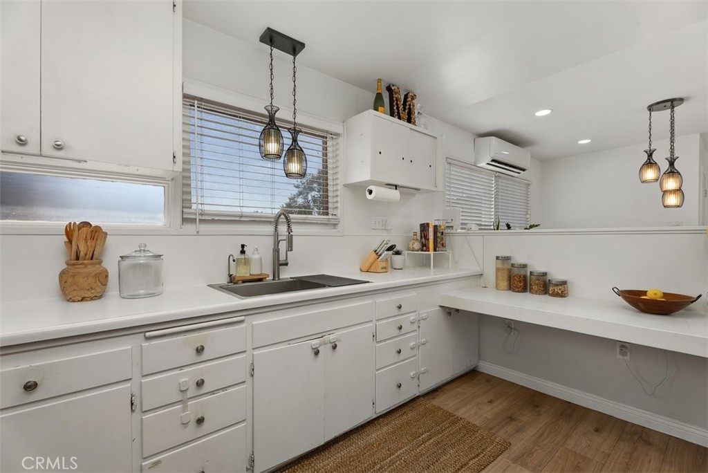 Bright white kitchen at 9925 Rutland Avenue with L-shaped counters, a stainless-steel sink beneath a window with blinds, pendant lights, and wood flooring.
