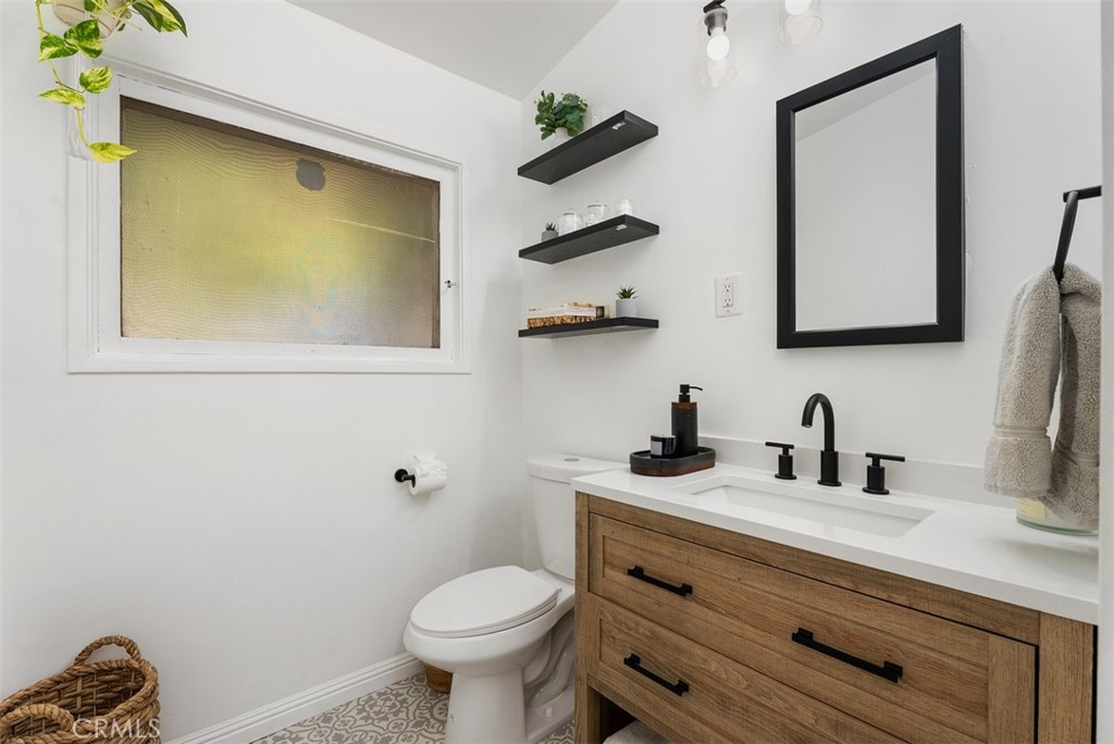Bathroom at 9925 Rutland Avenue with a wood vanity, white countertop, matte-black fixtures, toilet, frosted window, and floating shelves.