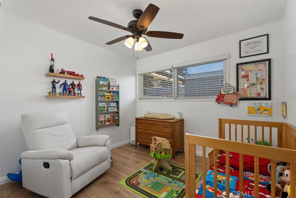 Bright nursery with a white recliner, wooden crib, toy shelves, dresser, and colorful play rug beneath a ceiling fan.