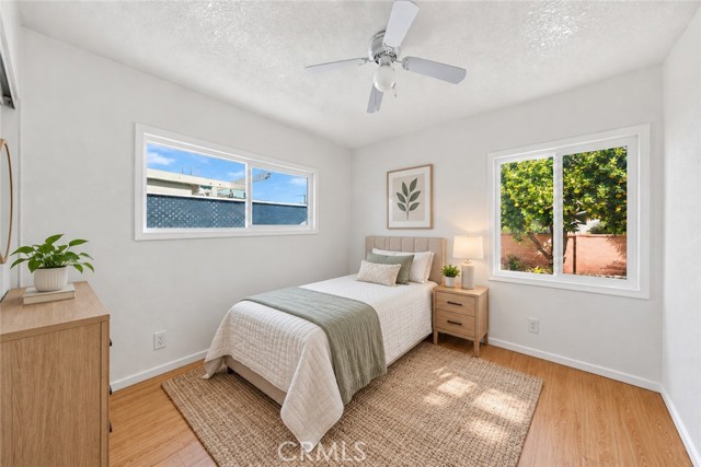 A well-lit bedroom with a twin bed, neutral-toned bedding, a wooden nightstand with a lamp, a potted plant on a dresser, and two windows offering views of the exterior.