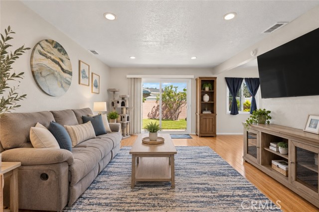 A furnished living room featuring a grey sectional sofa, a coffee table on a blue and white striped rug, a wall-mounted TV, and a sliding glass door leading to a backyard.