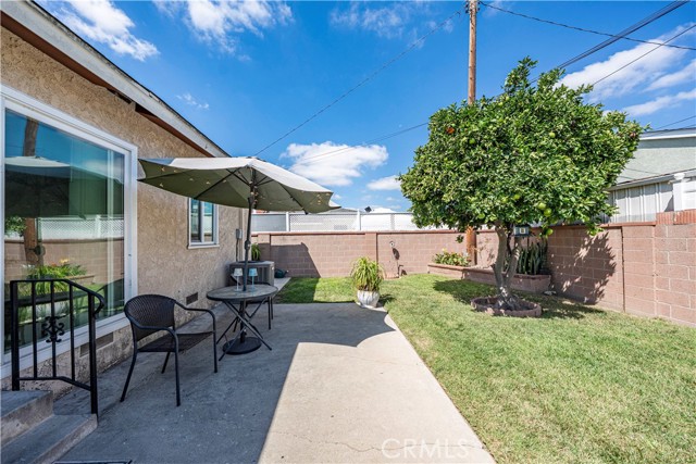 A small backyard patio featuring a dining set with a large umbrella, adjacent to a grassy area with a fruit tree and surrounded by a masonry block wall under a blue sky.