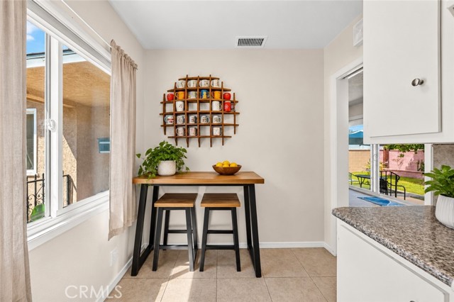 A small breakfast area featuring a high wooden table with two bar stools, a round wall-mounted mug rack, a plant, and a bowl of lemons, with a window and a doorway leading to the outdoors.