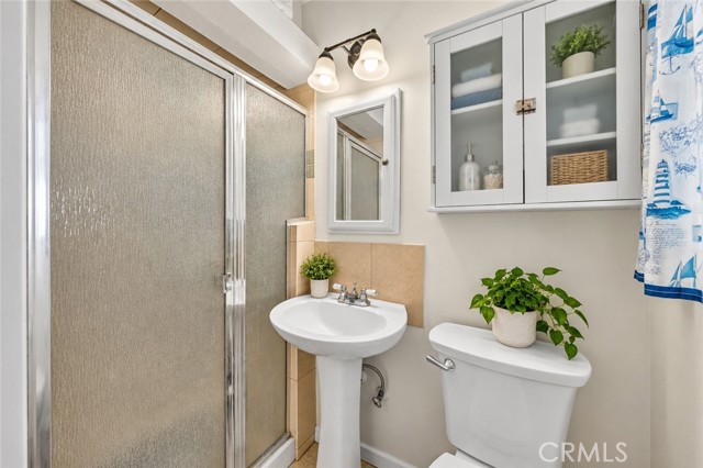 A small bathroom featuring a pedestal sink, toilet, glass-enclosed shower, and wall-mounted storage cabinet with nautical-themed decor.