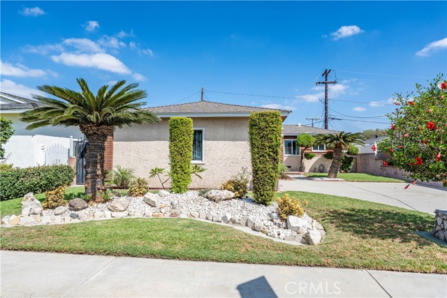 A front exterior view of a single-story beige stucco house with a palm tree, manicured hedges, and a gravel rock garden.