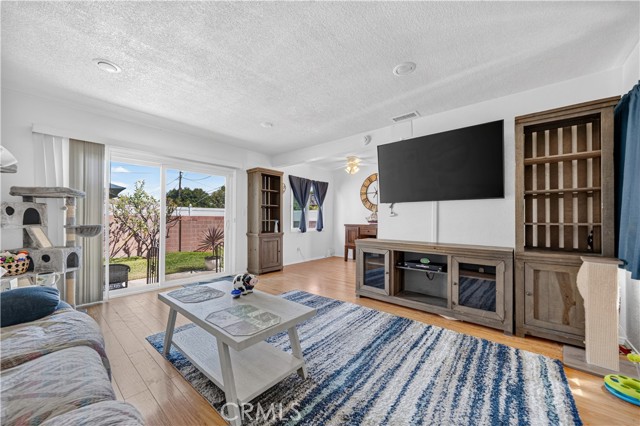 Spacious living room with light wood flooring, a large flat-screen TV mounted on the wall, a coffee table, and a sliding glass door looking out to a backyard.