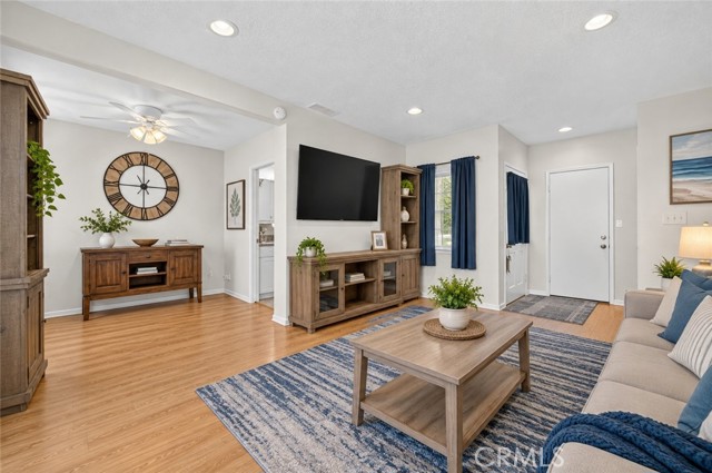 A furnished living room with light wood flooring, a blue rug, a large wall-mounted TV on a media console, and a dining area in the background with a circular wall clock.