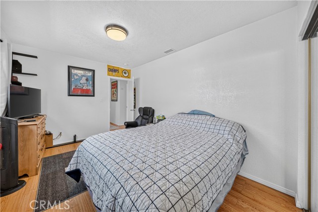 A bedroom with a queen sized bed, wood flooring, a flat screen TV on a dresser, and a small office space visible through the doorway.