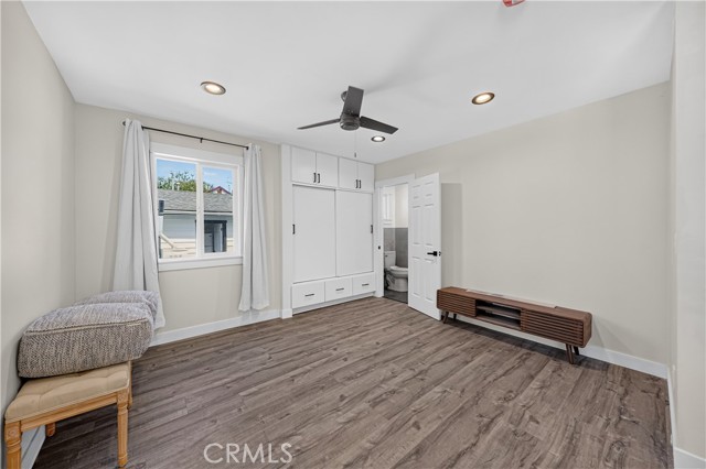 A bedroom with light gray walls, dark wood-look vinyl flooring, white built-in storage cabinets, a ceiling fan, and a low profile media console.