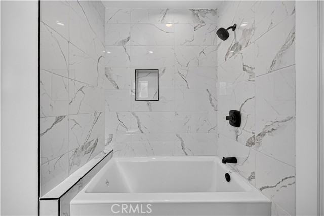 A modern bathroom featuring a white soaking tub with a black faucet against a white marble-look tiled wall with a built-in recessed niche and a black shower head.