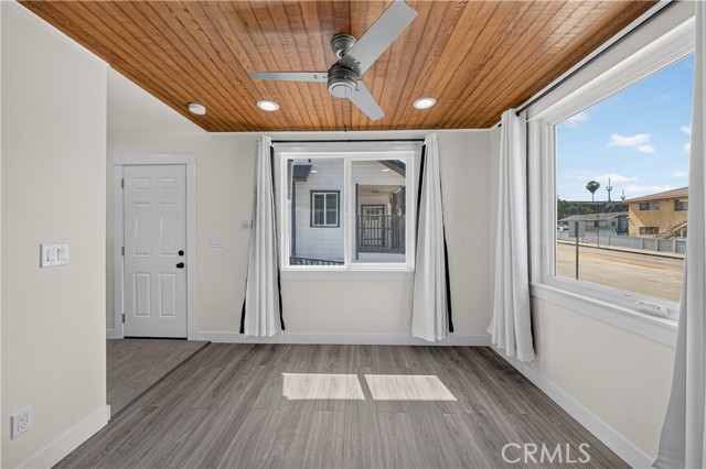 A brightly lit living room with wood plank ceiling, light gray wood-look flooring, and white walls, looking out through large windows.