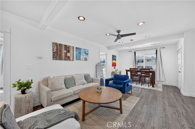 A bright living room with a beige sofa, a blue accent chair, a round wooden coffee table, and a dining set in the background, featuring grey laminate flooring and white walls.