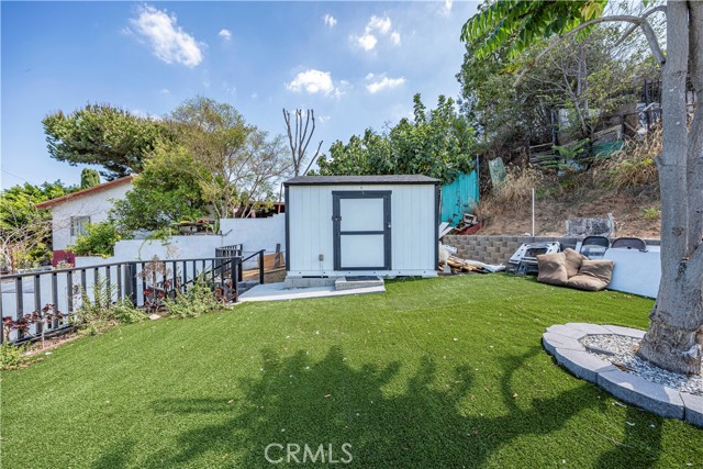 A backyard featuring a small storage shed, artificial grass lawn, retaining wall, and mature trees under a blue sky.