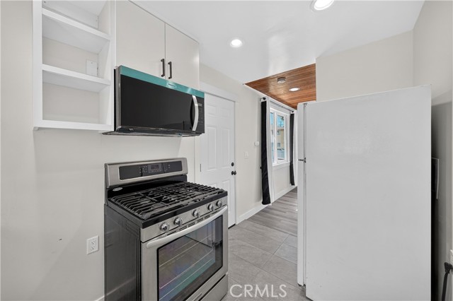 Modern kitchen with white cabinets, stainless steel gas stove, microwave, and white refrigerator, with tile flooring and a view into a hallway with wood-paneled ceiling.
