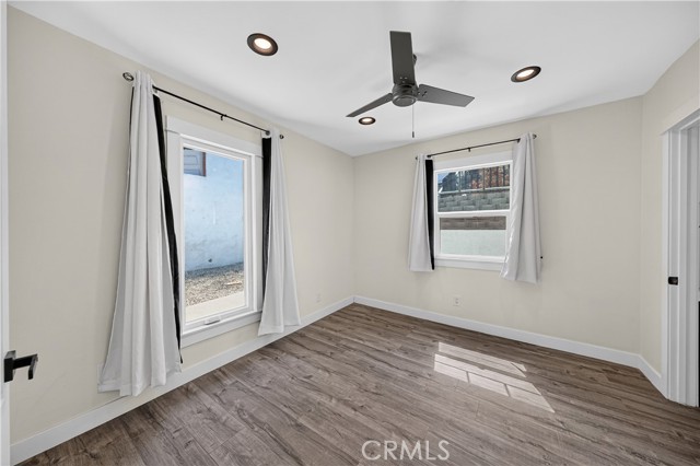 A vacant bedroom with beige walls, wood-look flooring, two windows with white curtains, recessed lighting, and a modern ceiling fan.