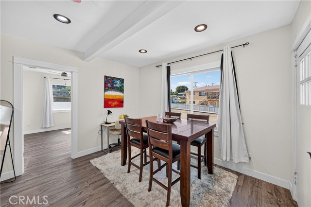 A well-lit dining room featuring a wooden table with four high-back chairs sitting on a shaggy rug, with white walls and a view into an adjacent room.