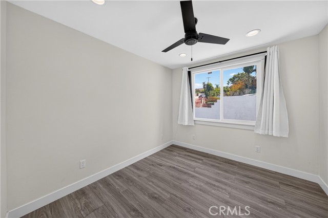 A vacant bedroom with light-colored walls, wood-look plank flooring, a black ceiling fan, and a single window with white curtains.
