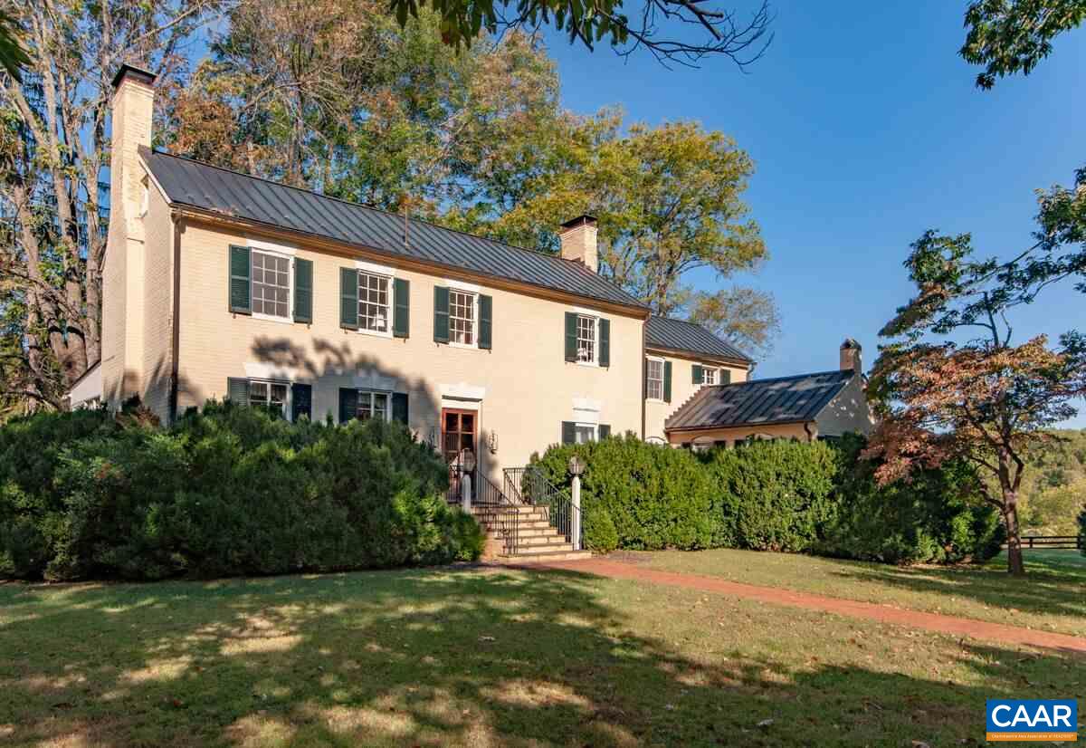 Pale brick house with dark metal roofs and green shutters, surrounded by mature trees.