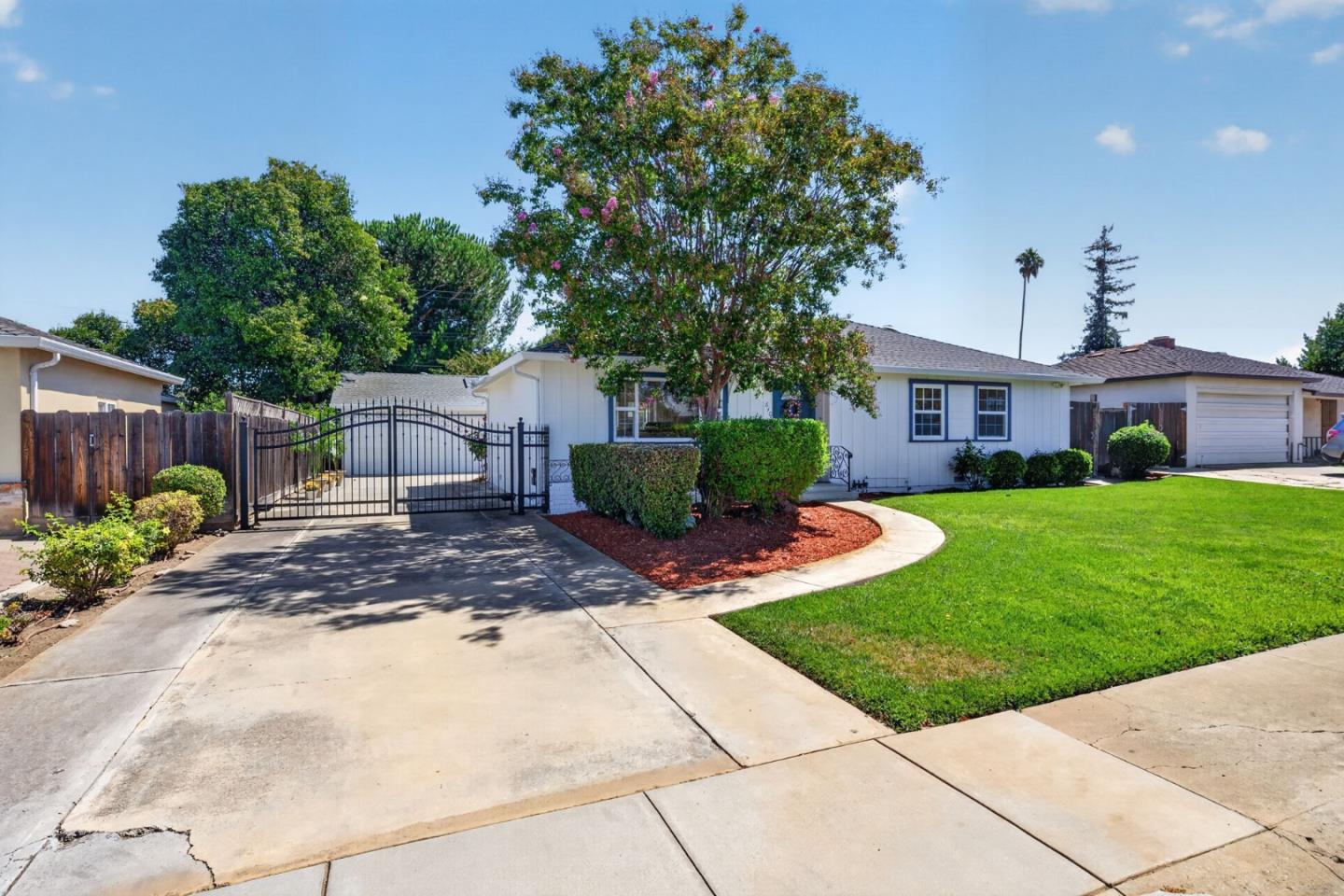 Single-story white house with a gated driveway, broad green lawn, and flowering tree.