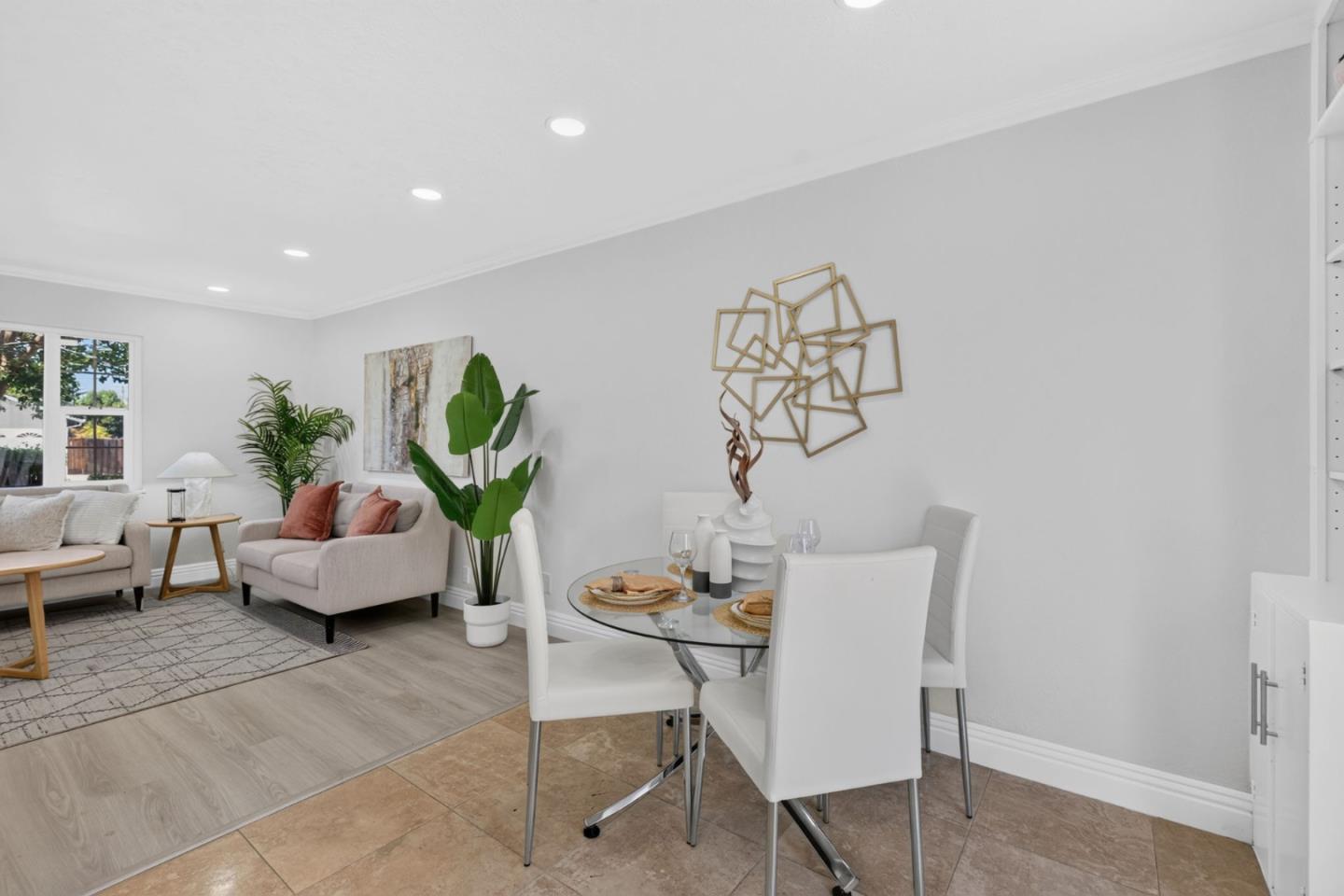 Staged living and dining room with gray sofas, a glass table, white chairs, and tropical plants.