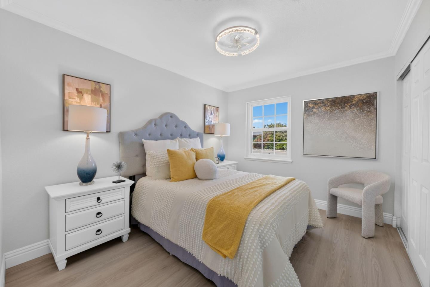 Staged bedroom with a tufted gray headboard, mustard accents, and a sunlit window.