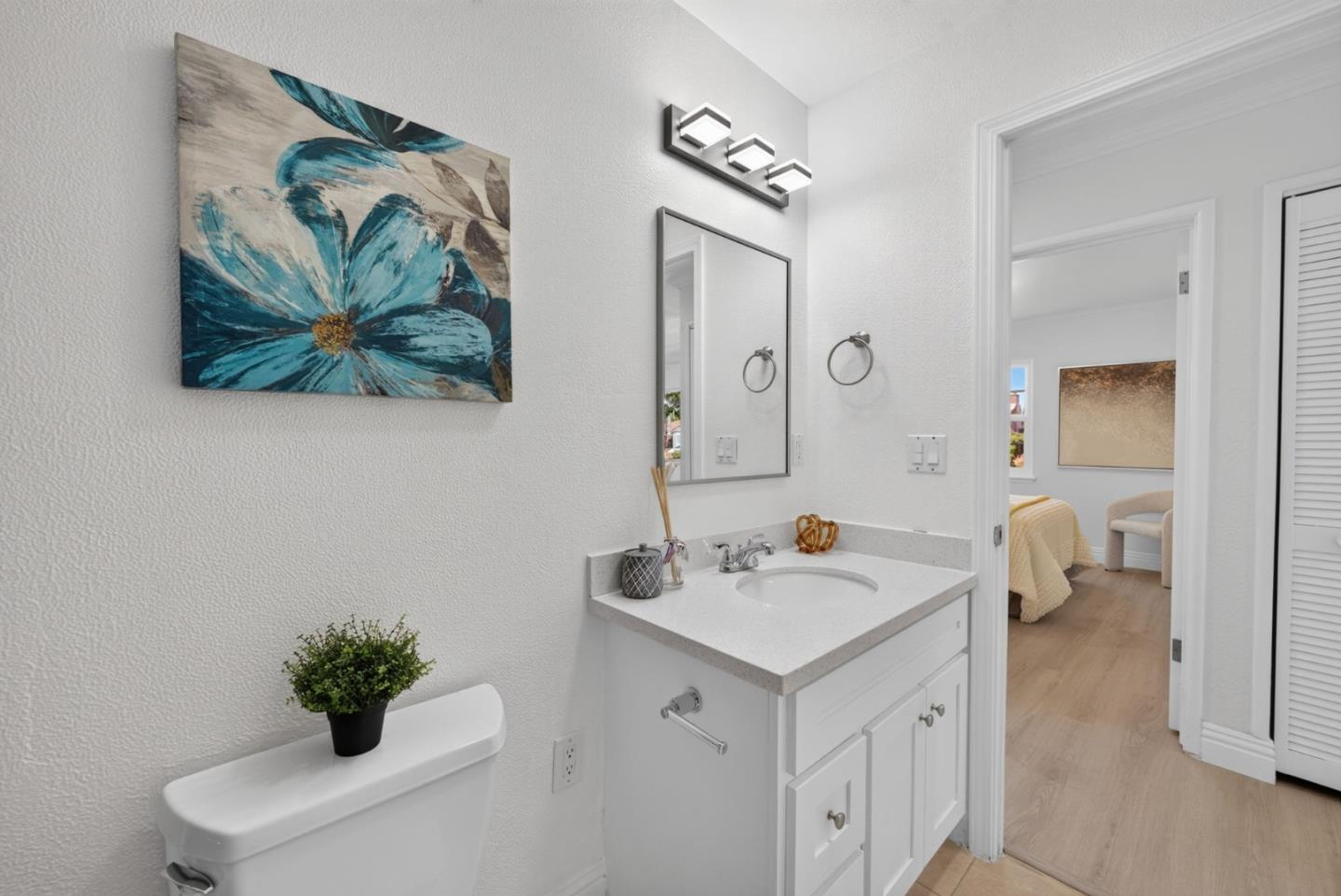 Bathroom with white vanity and toilet, blue floral wall art, and an open doorway to a bedroom.