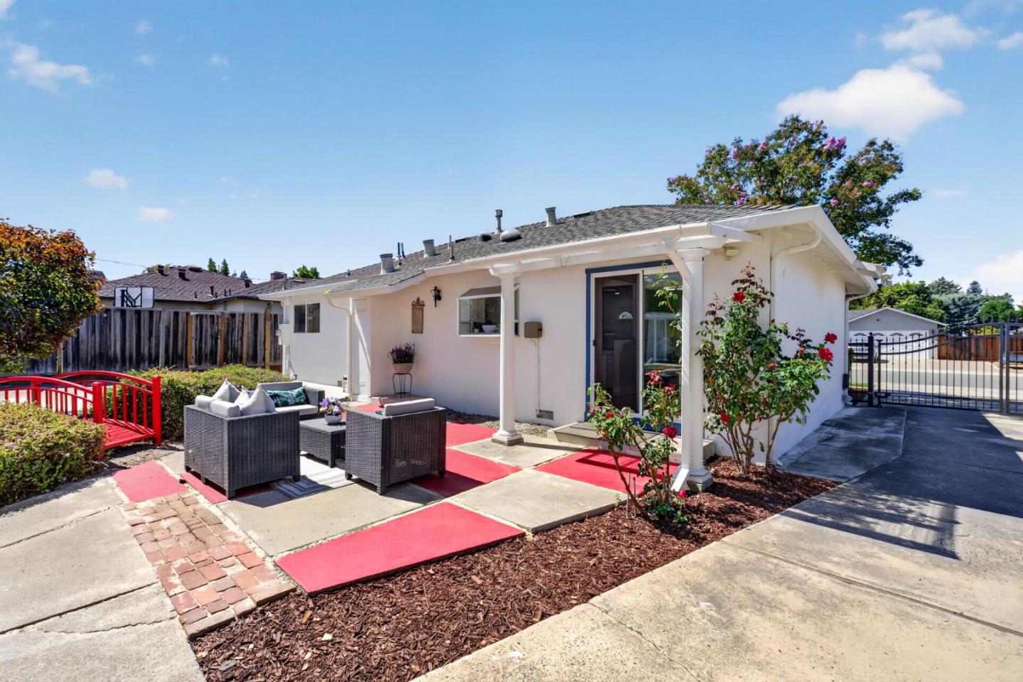 White home with a furnished patio, red garden bridge, and flowering rose bushes.