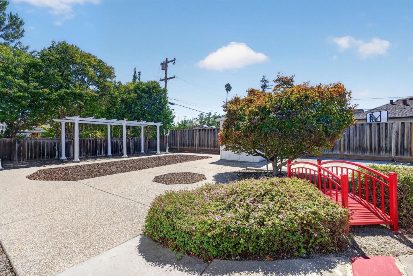 Backyard patio with a white pergola, flowering shrubs, and a bright red footbridge.