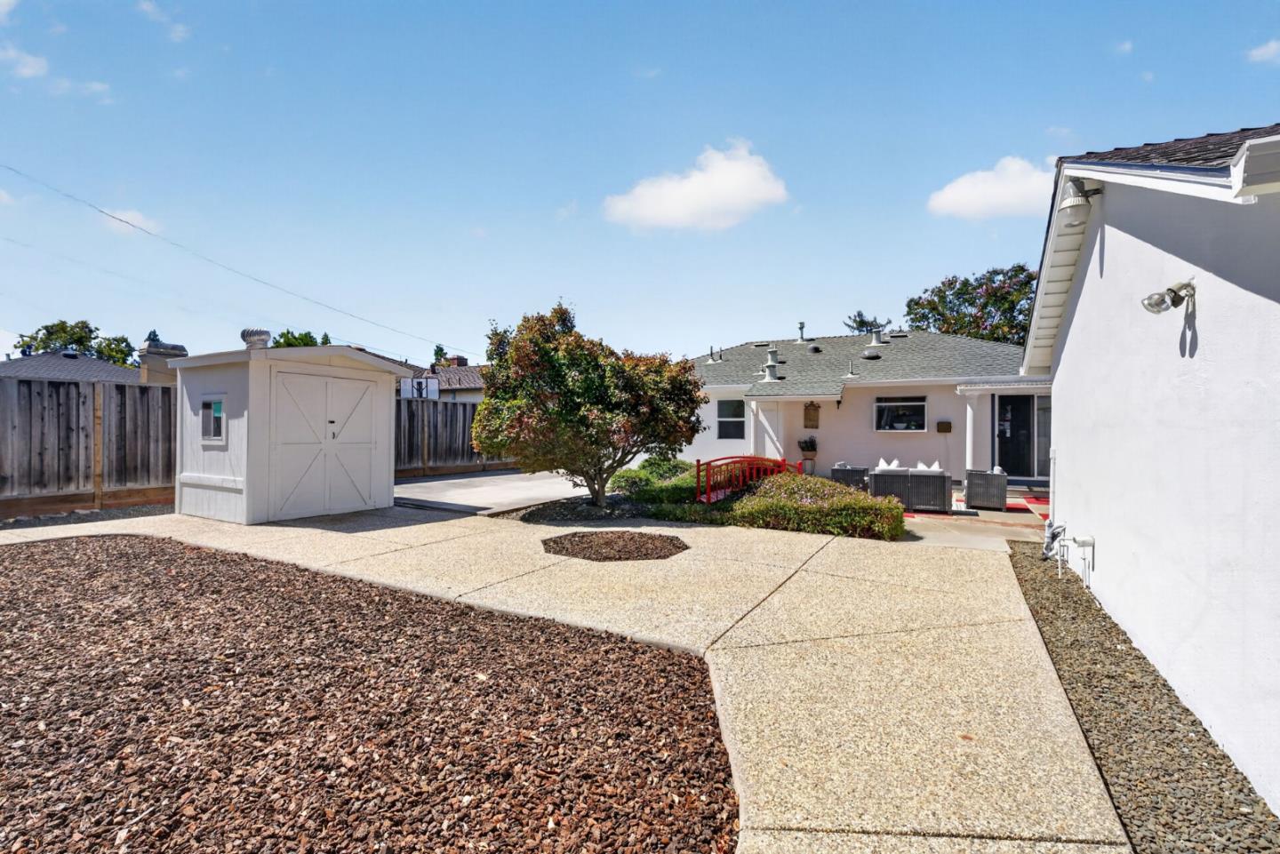 Backyard with a white shed, concrete walkway, landscaped tree, red garden bridge, and patio seating.