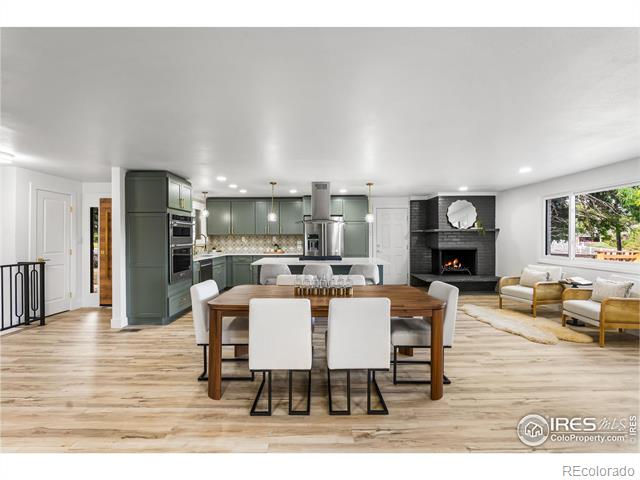 Open-plan kitchen and dining area with gray-green cabinets, pale wood floors, and a brick fireplace.
